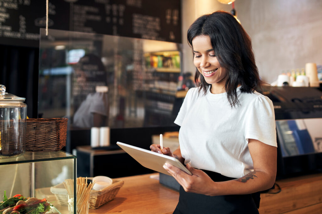Restaurant franchise owner smiling while using a tablet behind the counter of a café, reviewing orders and managing daily business operations.