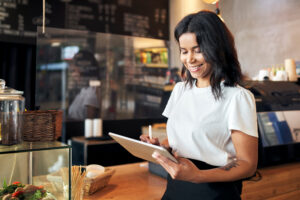 Restaurant franchise owner smiling while using a tablet behind the counter of a café, reviewing orders and managing daily business operations.